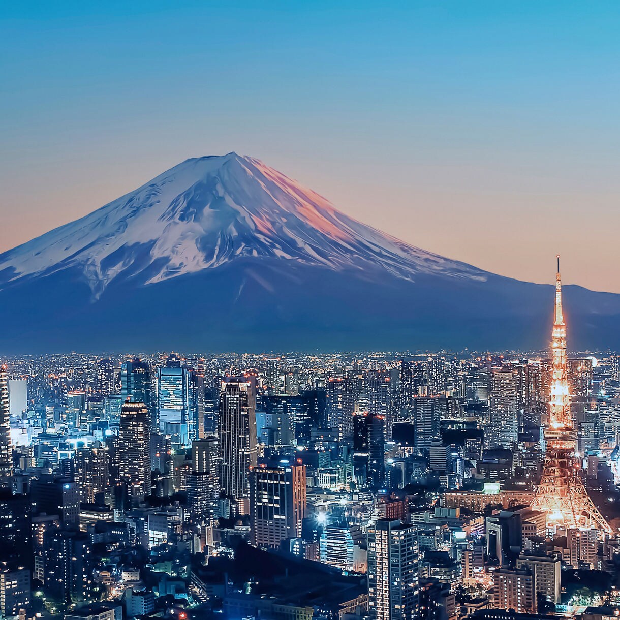 Aerial twilight view of Tokyo's illuminated skyline with the glowing Tokyo Tower prominent among densely packed skyscrapers and the snow-capped peak of Mount Fuji rising majestically against a blue and orange gradient sky in the background.