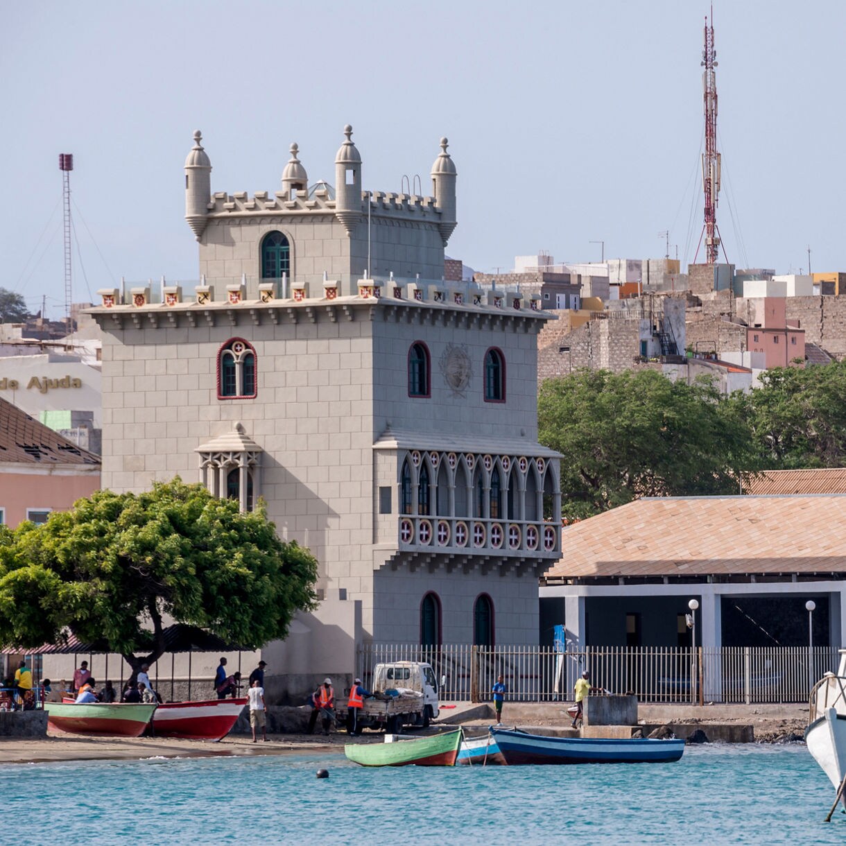 Coastal scene in Mindelo featuring a gray, castle-like tower with arched windows, small fishing boats pulled to shore and pastel buildings rising behind it under a clear sky.
