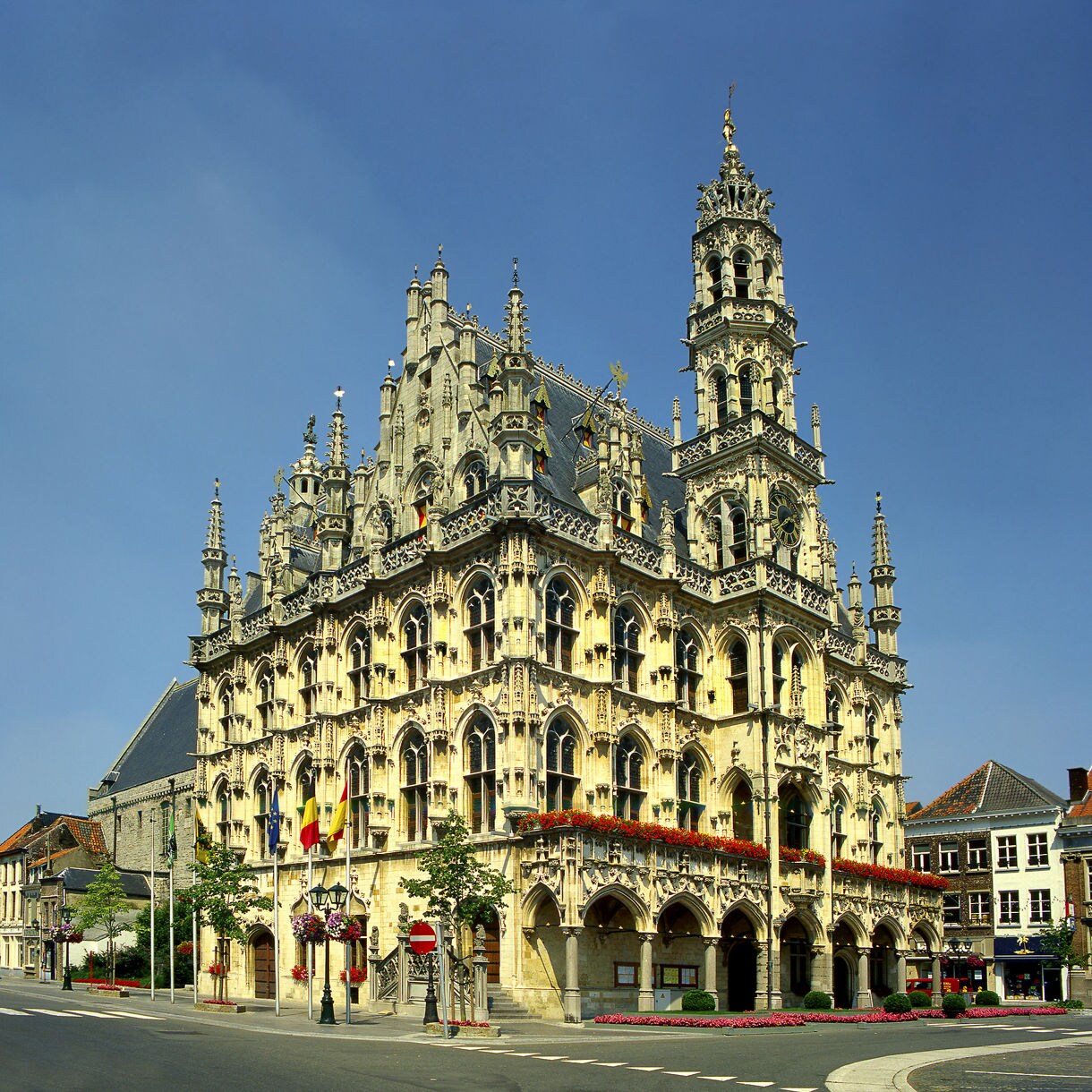Exterior view of the ornate Gothic-style Town Hall in Leuven, Belgium, featuring intricate stone carvings, tall spires, arched windows and flags along the facade.