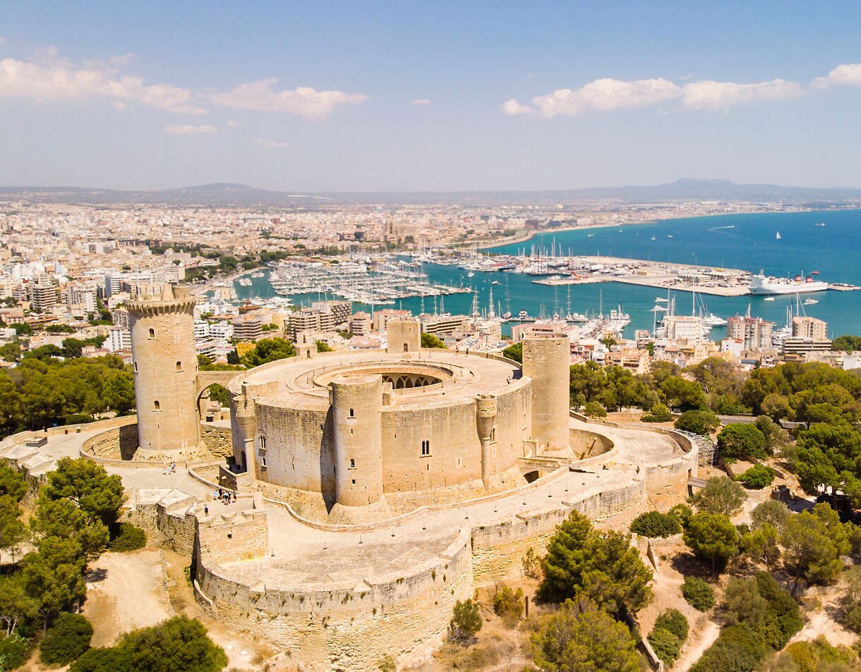 Aerial view of Bellver Castle in Palma de Mallorca, a circular medieval fortress surrounded by pine trees with the city and marina in the background.
