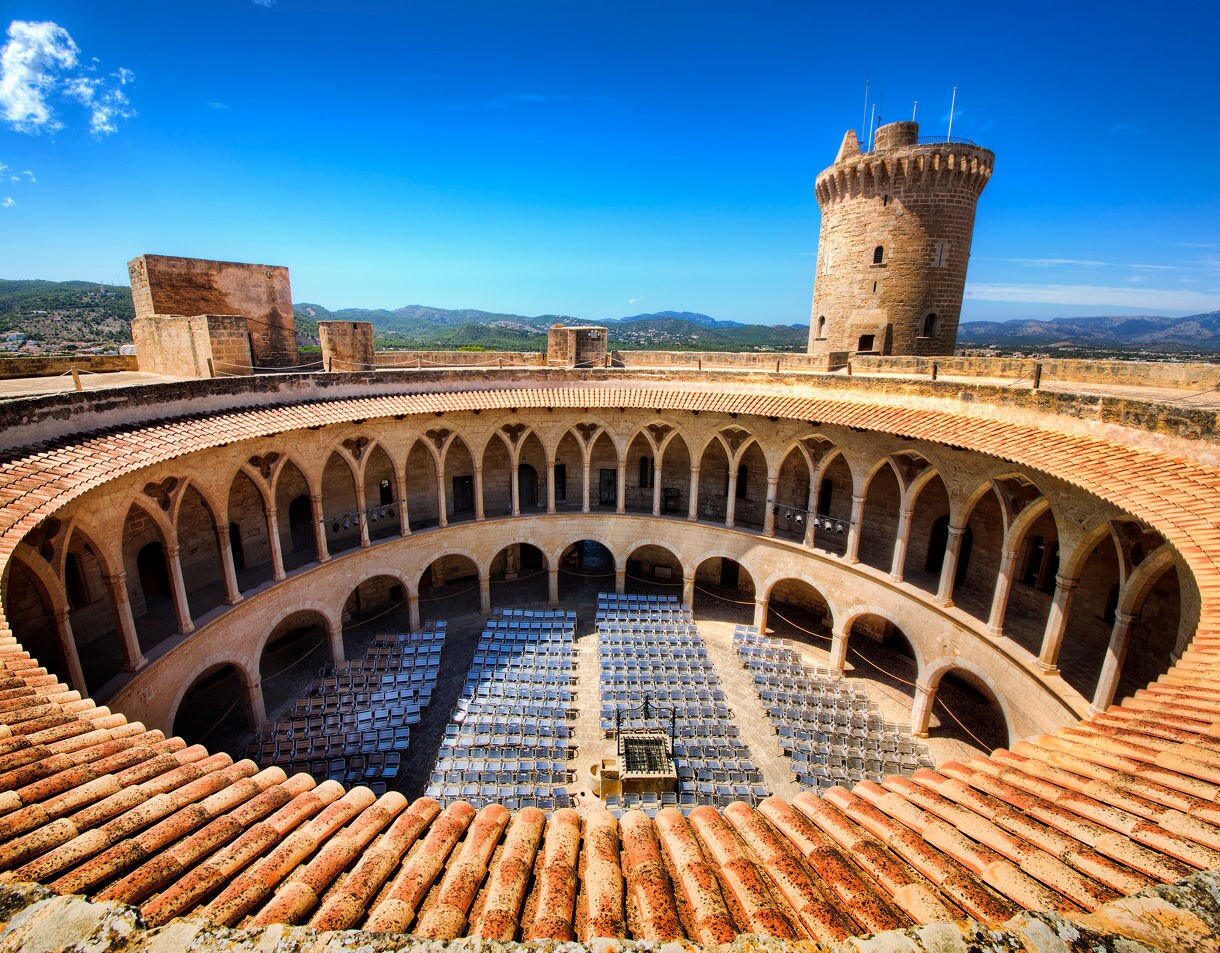 Interior view of Bellver Castle in Palma, Mallorca, featuring a round courtyard lined with Gothic arches and a tall stone tower under a clear blue sky.