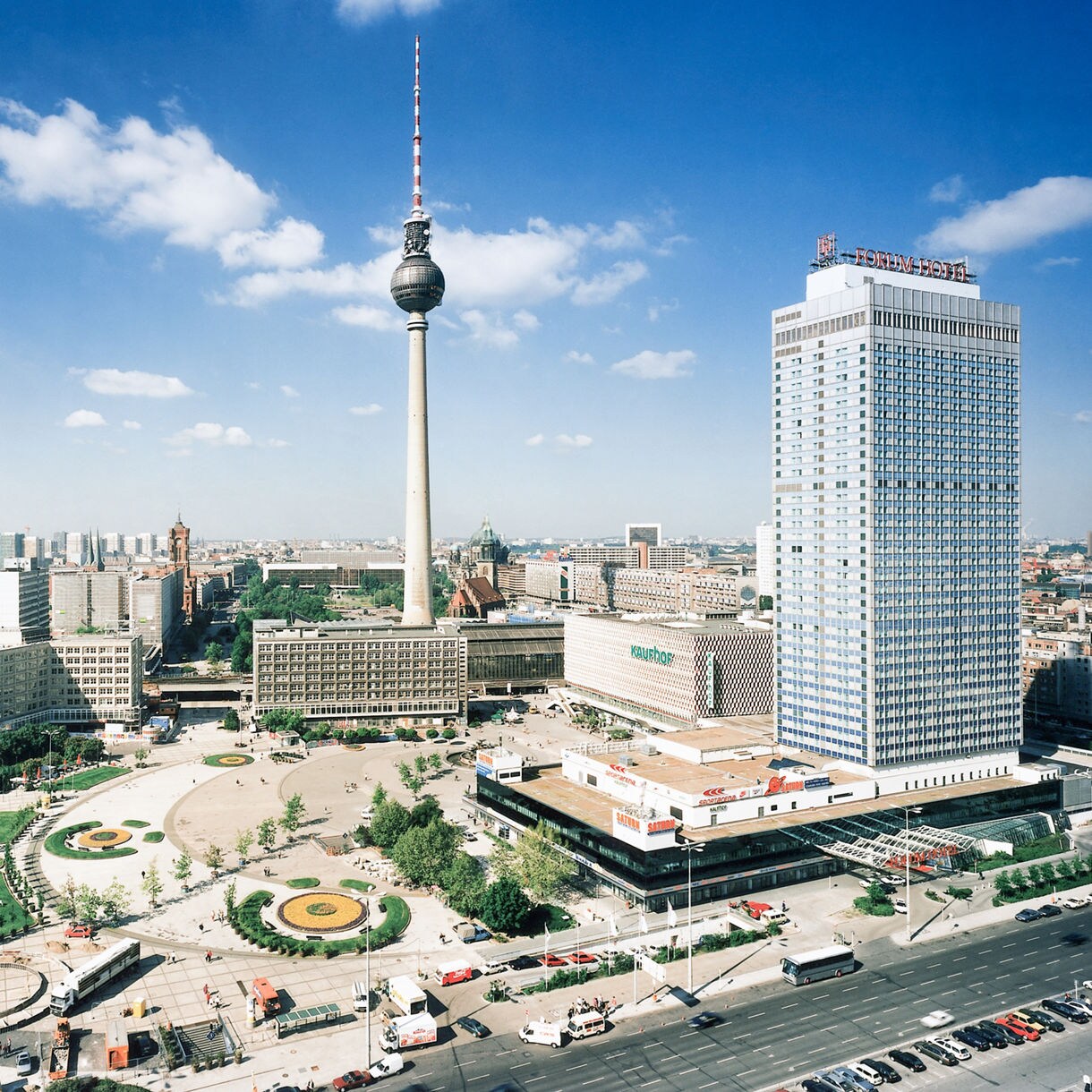 Aerial view of Berlin’s Alexanderplatz with the Fernsehturm TV Tower, modern high-rises, open square and busy streets under a bright blue sky.
