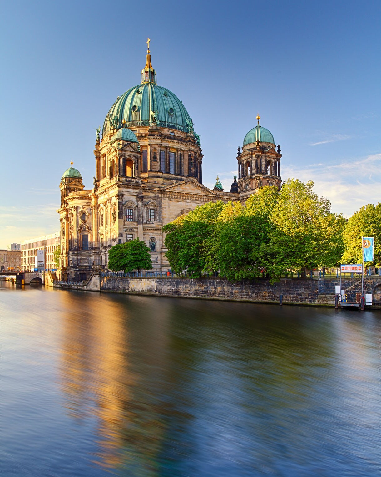 Berlin Cathedral with its green copper dome and ornate towers reflected in the calm waters of the River Spree on a clear evening.