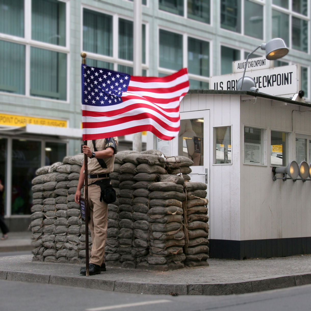 Guard post at Berlin’s Checkpoint Charlie with sandbags, a small white hut, an American flag and surrounding city buildings.