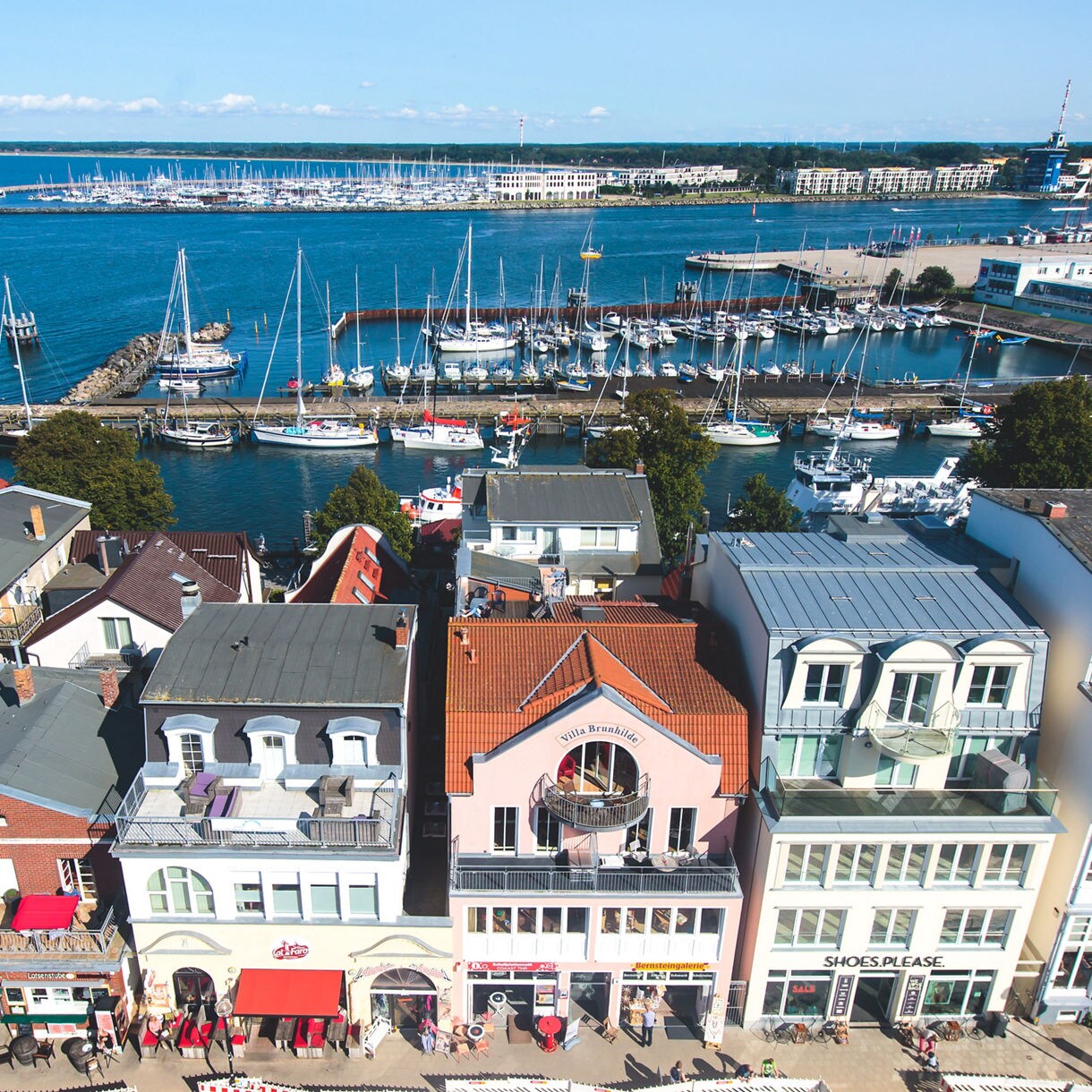 Aerial view of Warnemünde, Germany with pastel waterfront houses, outdoor cafes and a harbor full of sailboats along the Baltic coast.