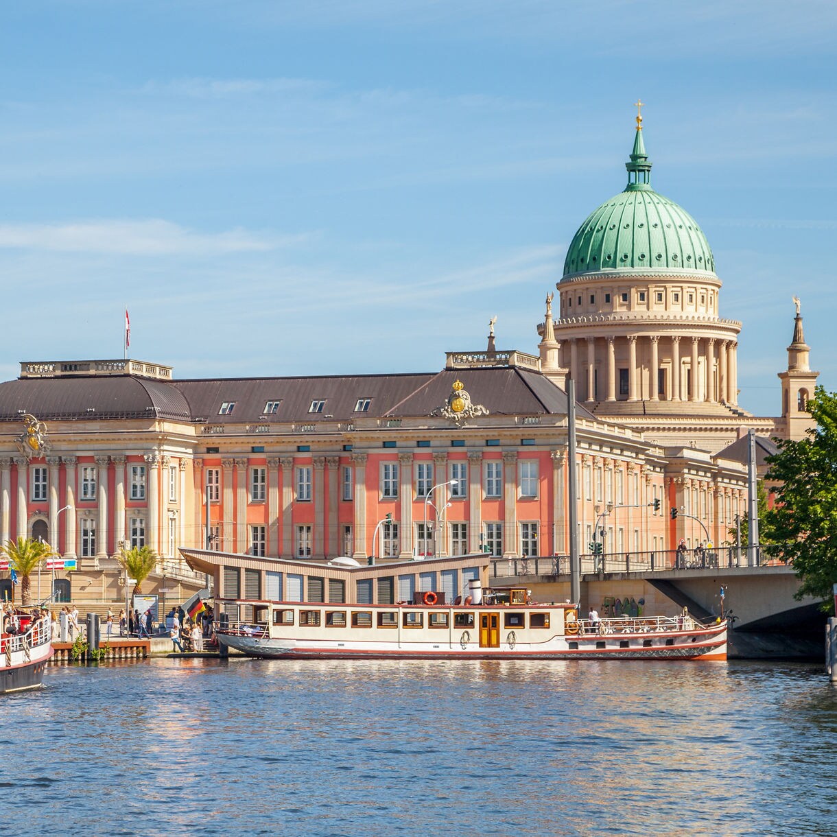 View of Potsdam’s Old Market Square with the pink baroque City Palace and St. Nicholas’ Church dome behind, seen from the river with boats docked in front.