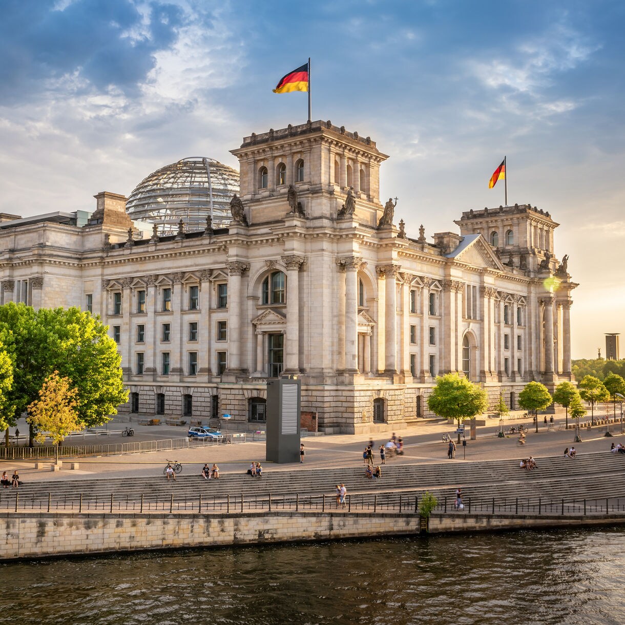 Exterior of the Reichstag building in Berlin with German flags flying, stone façade and the glass dome visible behind viewed from across the river.