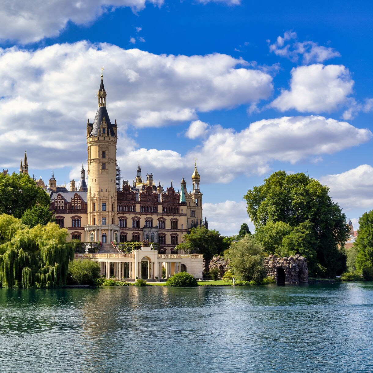 Schwerin Castle in Germany with ornate towers and golden domes surrounded by trees and reflected in the calm blue lake below.