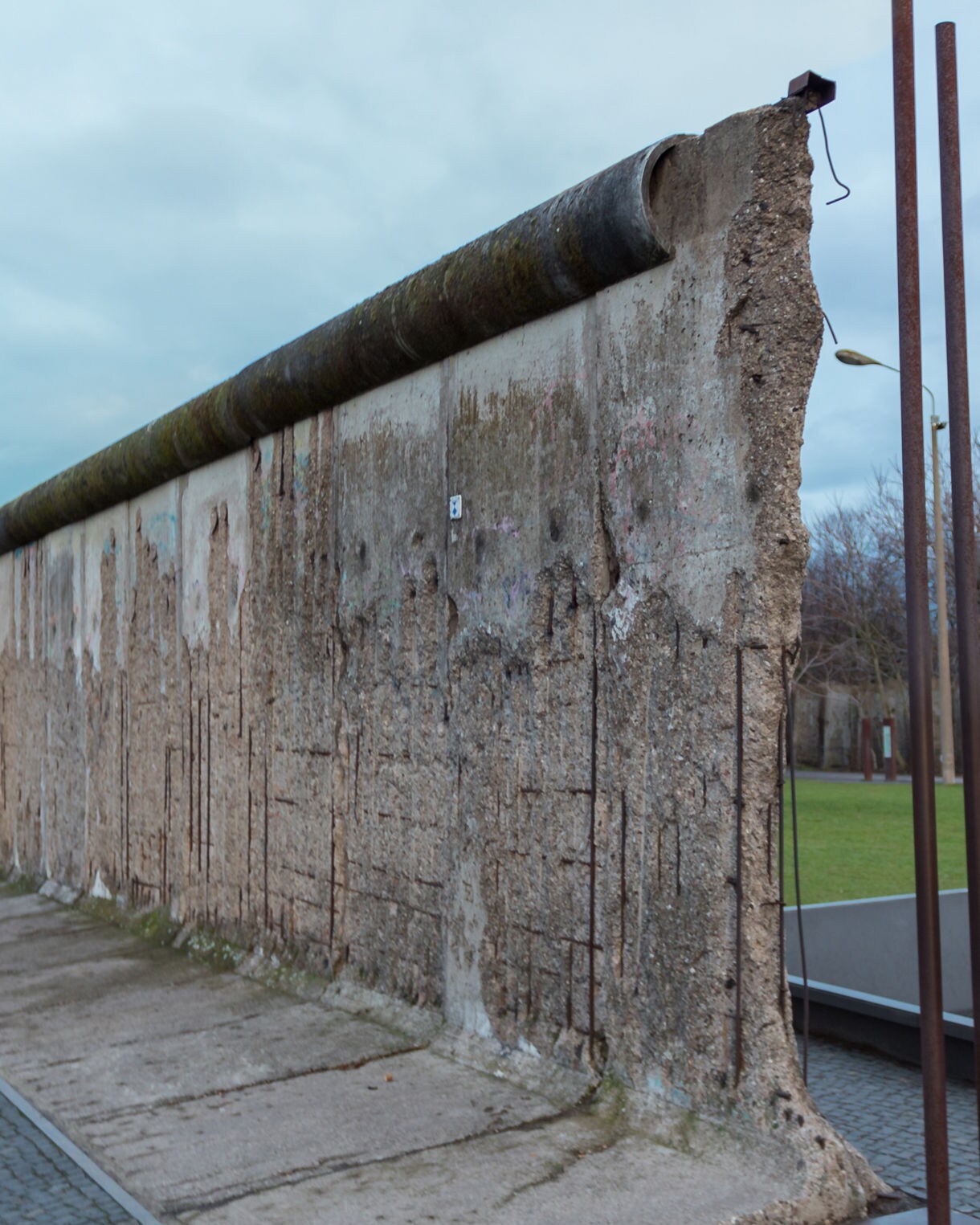 A preserved section of the Berlin Wall showing weathered concrete with exposed rebar, set along a pathway with grass and city buildings in the background.