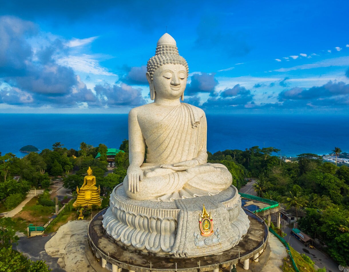 Aerial view of the massive white Big Buddha statue on a hilltop in Phuket with ocean and city views in the background.