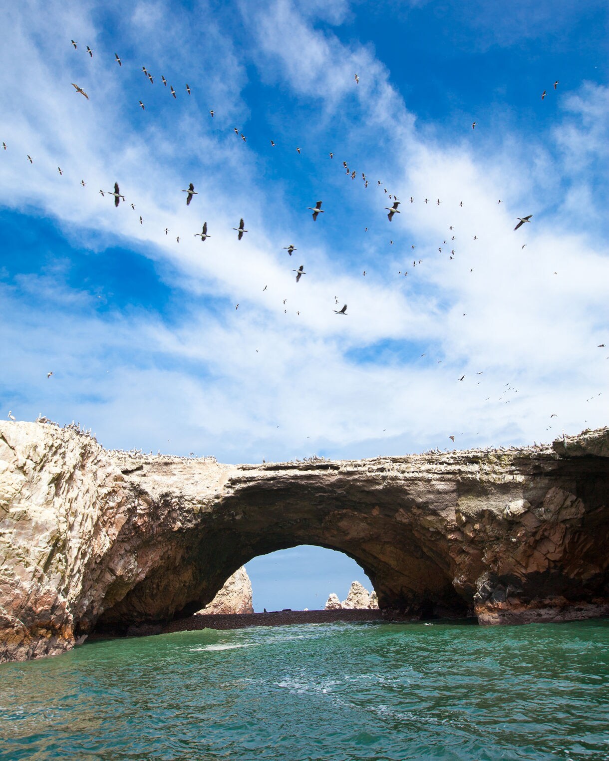 Rocky sea arch at the Ballestas Islands with green water below and flocks of seabirds flying overhead against a bright blue sky.