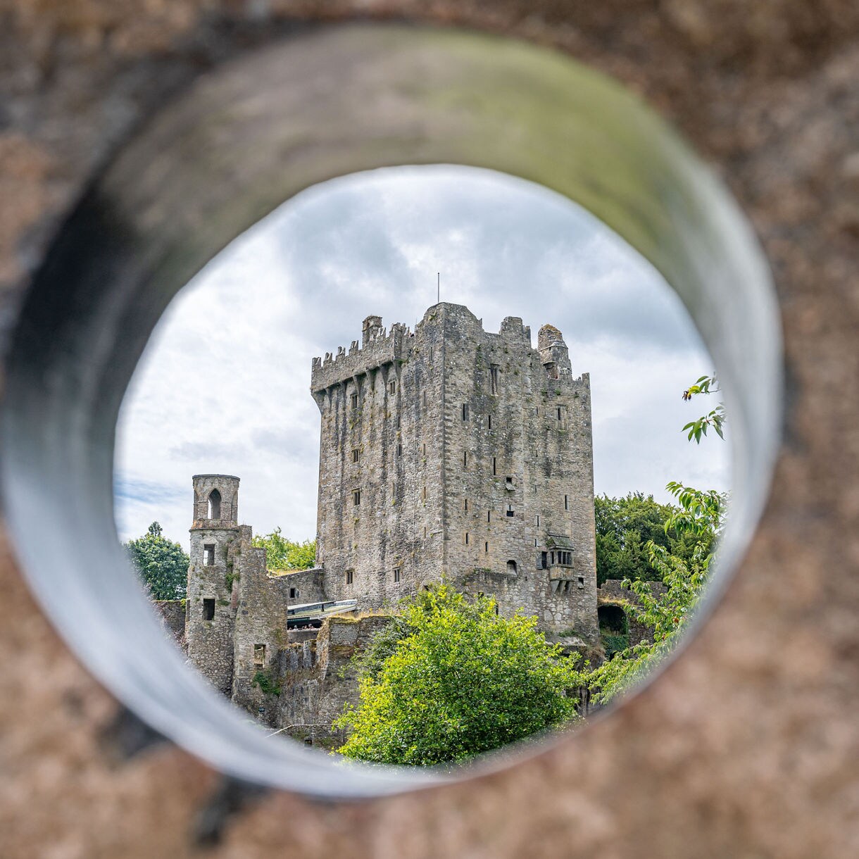 Blarney Castle in Ireland seen through a circular opening in stone, with its tall tower and surrounding greenery in clear focus against a cloudy sky.