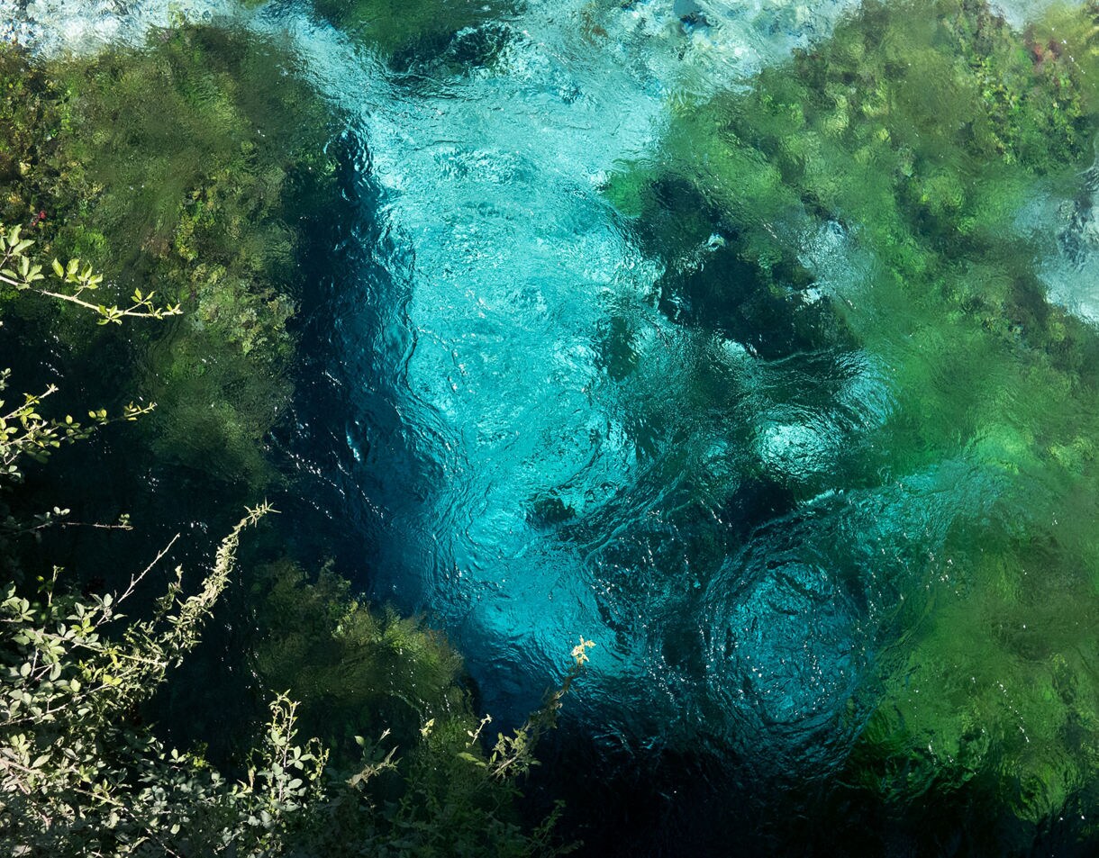 Overhead view of the Blue Eye spring in Albania, showing vibrant turquoise water swirling above deep underwater caverns surrounded by lush green aquatic plants.