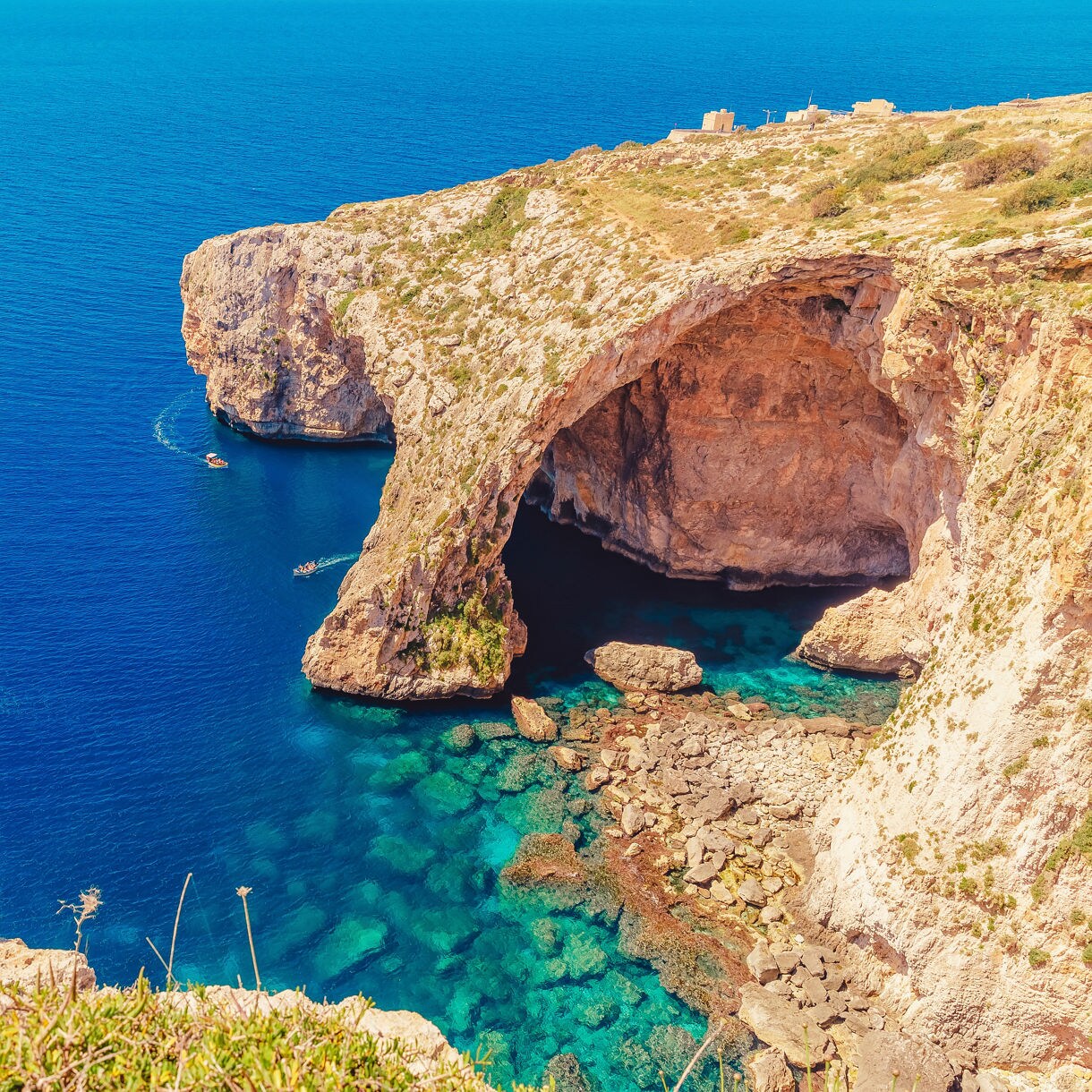 View of Malta’s Blue Grotto, showing a massive limestone arch over clear turquoise water with small boats near the cave entrance.