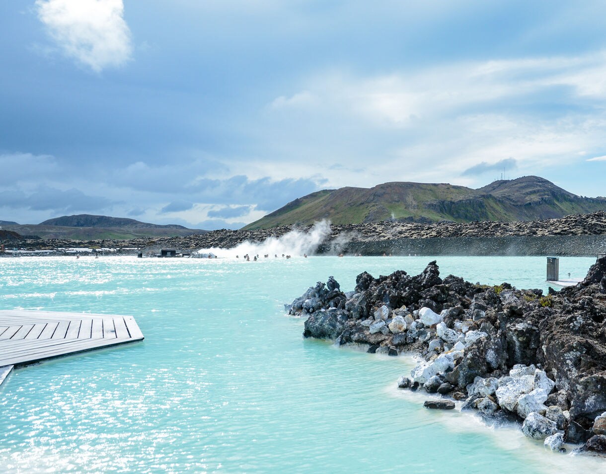 Geothermal Blue Lagoon in Iceland with steaming turquoise waters surrounded by dark volcanic rock and distant mountains.