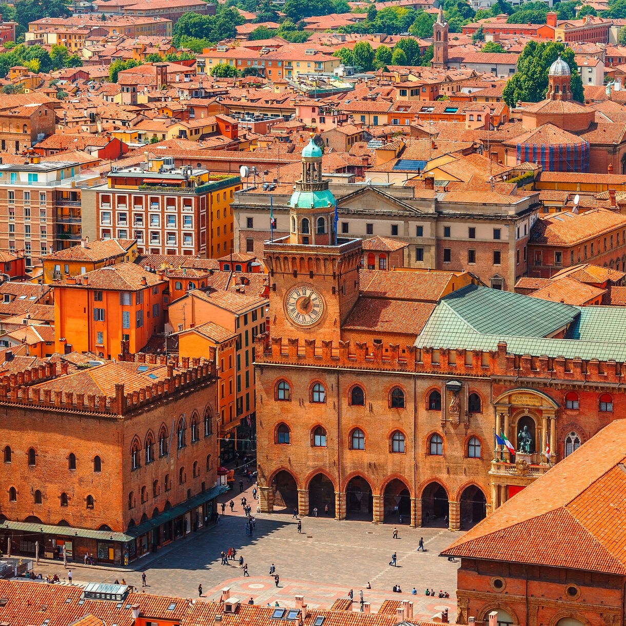 Panoramic view of Bologna featuring Piazza Maggiore, the Palazzo d'Accursio clock tower and terracotta rooftops stretching across the city.