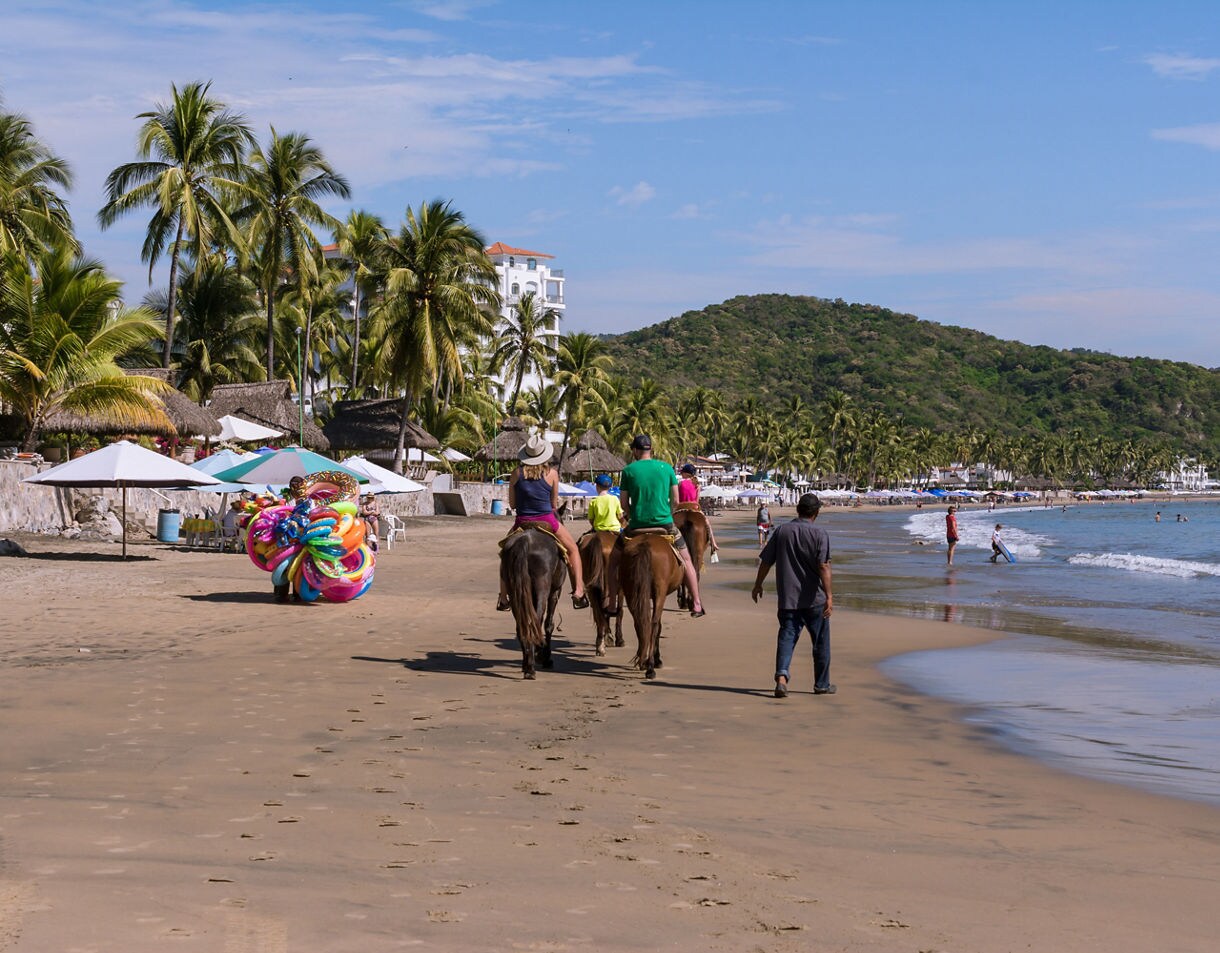 Visitors riding horses on La Boquita Beach with palm trees, umbrellas and calm ocean waves stretching toward green hills.