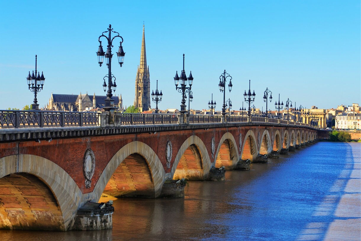 A long brick and stone arched bridge in Bordeaux lined with ornate lampposts, spanning a calm river with a tall church spire and historic buildings visible in the background under a clear blue sky.