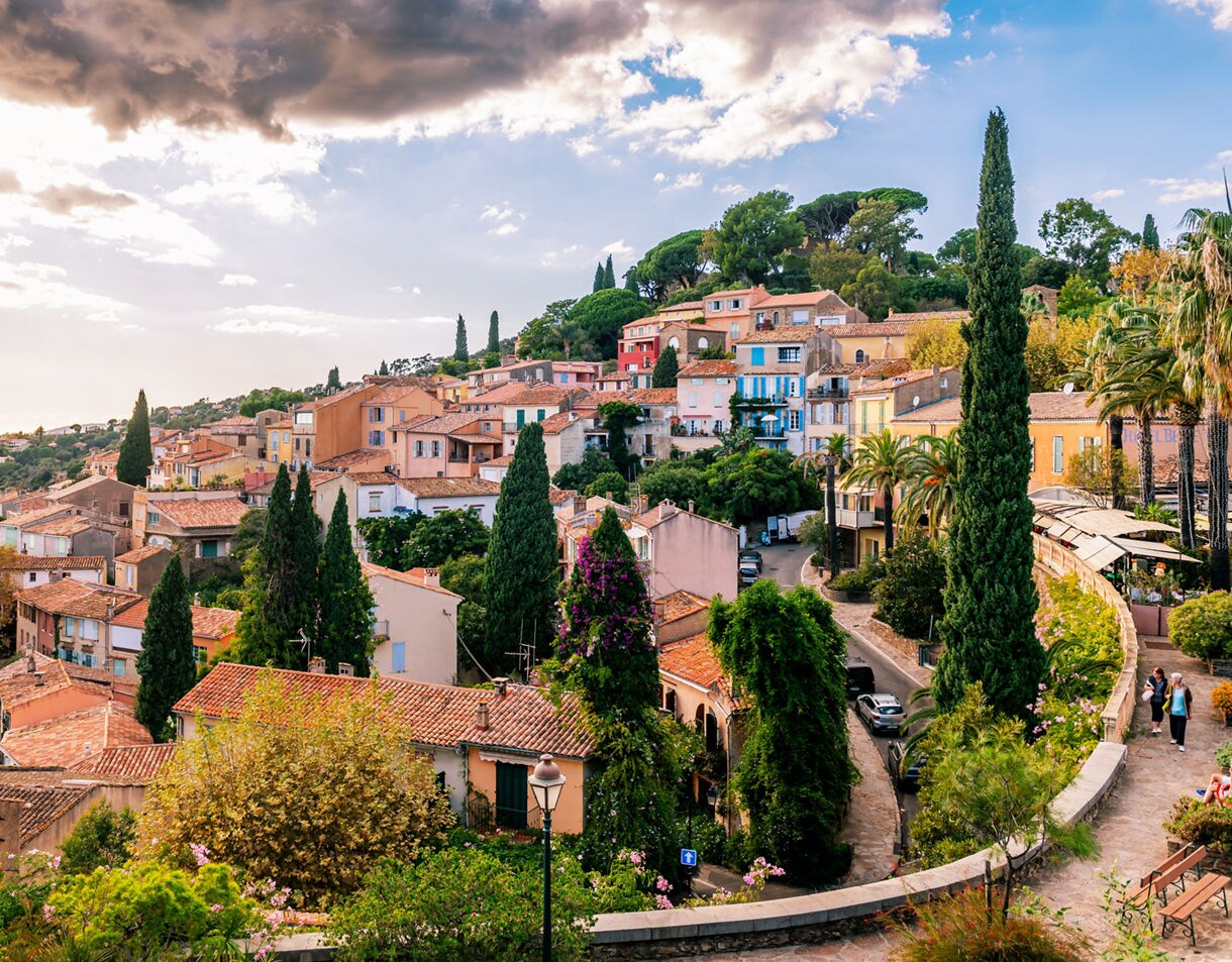 Elevated view of Bormes-les-Mimosas showing terracotta rooftops, pastel houses, tall cypress trees and winding village streets set against rolling hills and a partly clouded sky.