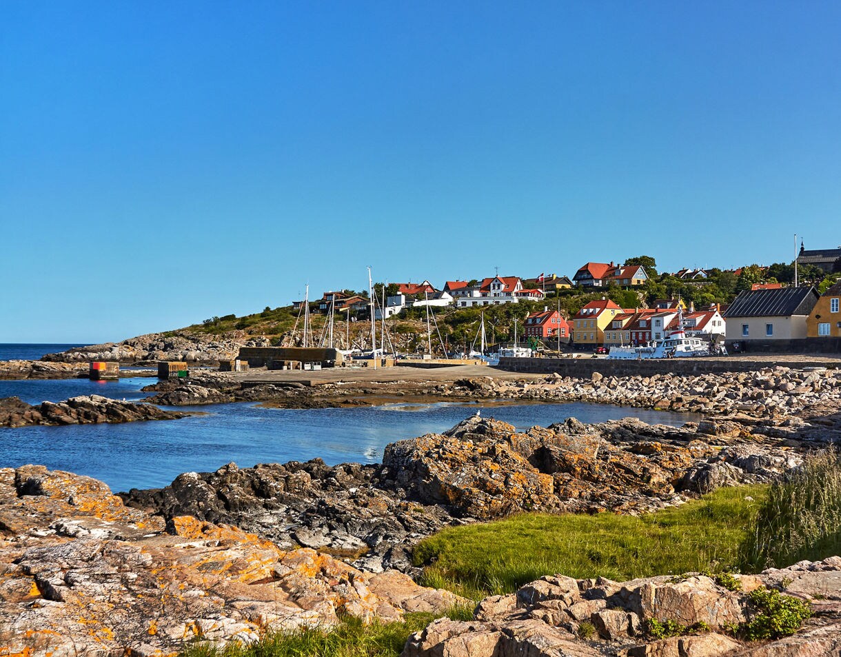 Coastal view of Gudhjem on Bornholm, showing colorful houses with red roofs perched on a hillside, sailboats in the harbor, rocky shoreline in the foreground and a clear blue sky overhead.