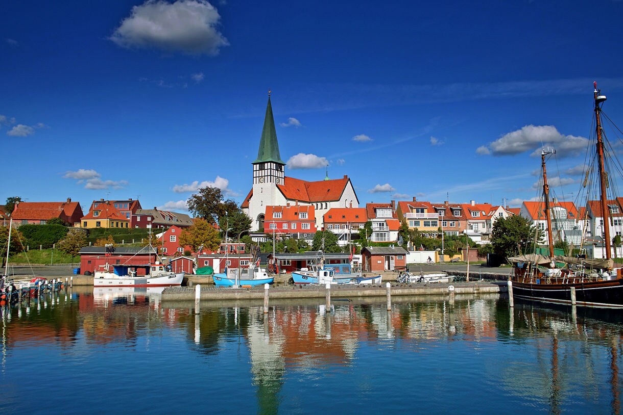 A vibrant harbor scene in Bornholm with small boats docked on calm blue water, colorful houses lining the shore and a large white church with a tall green spire rising above the town.