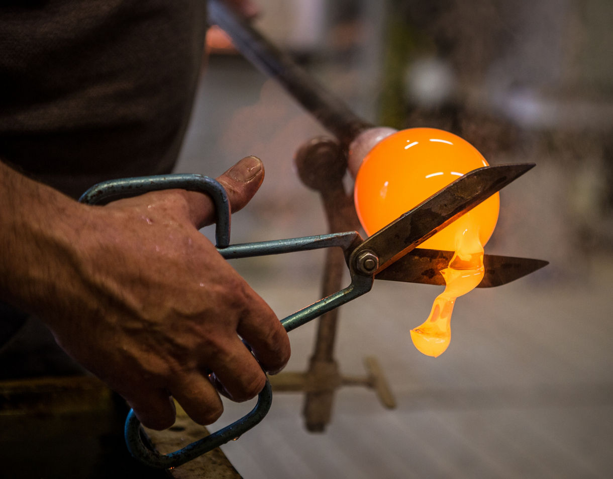 Close-up of a glassblower’s hands using metal shears to trim a glowing orange blob of molten glass at the end of a blowpipe inside a workshop.