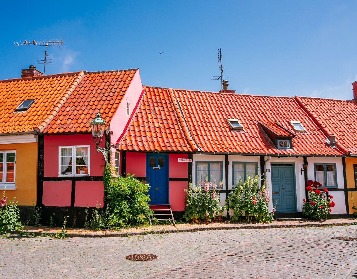 Row of colorful half-timbered houses in Rønne, Bornholm, featuring red tile roofs, pastel walls, flowers along the cobblestone street and a clear blue summer sky overhead.