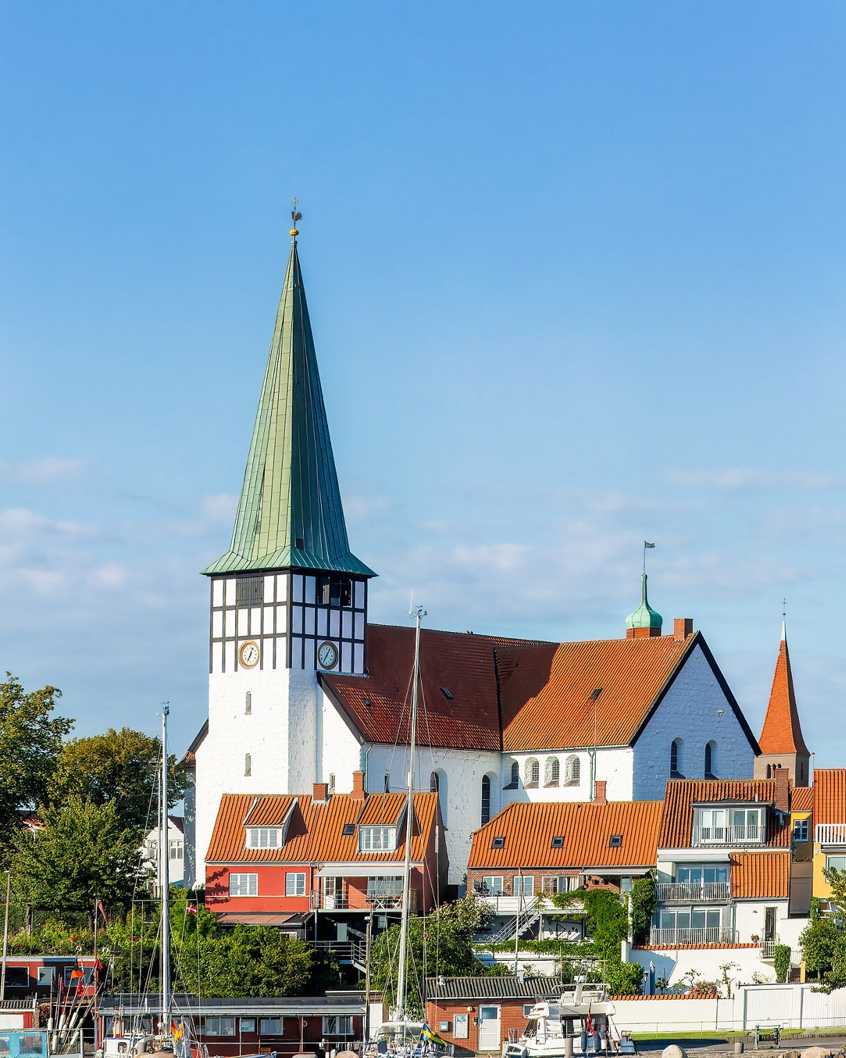 Tall white church with a green spire and red-tiled roof overlooking colorful houses and a small harbor on the island of Bornholm under a clear blue sky.