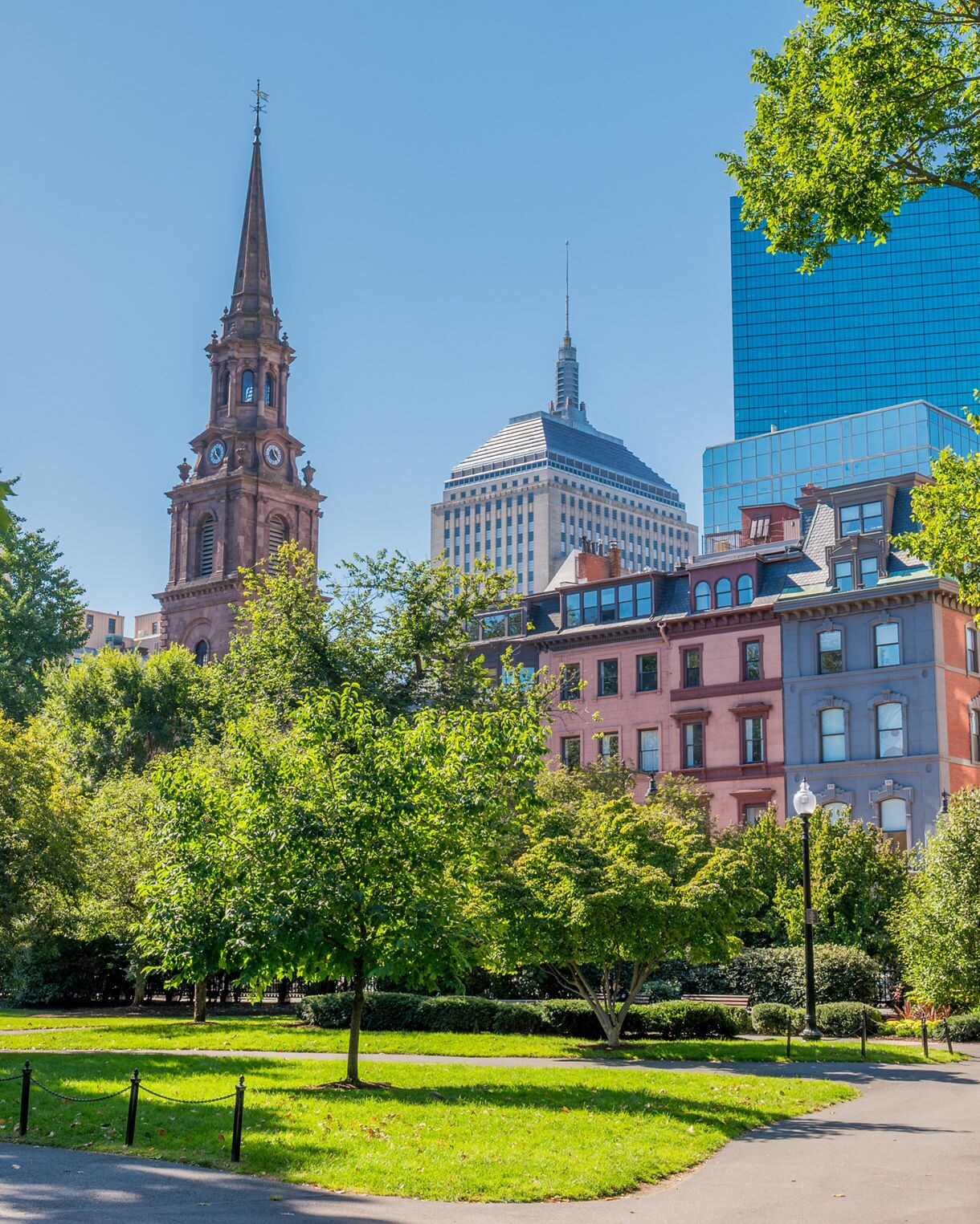 Boston Common with winding paths, lush summer trees and views of historic brick buildings and a tall church spire against a clear blue sky.