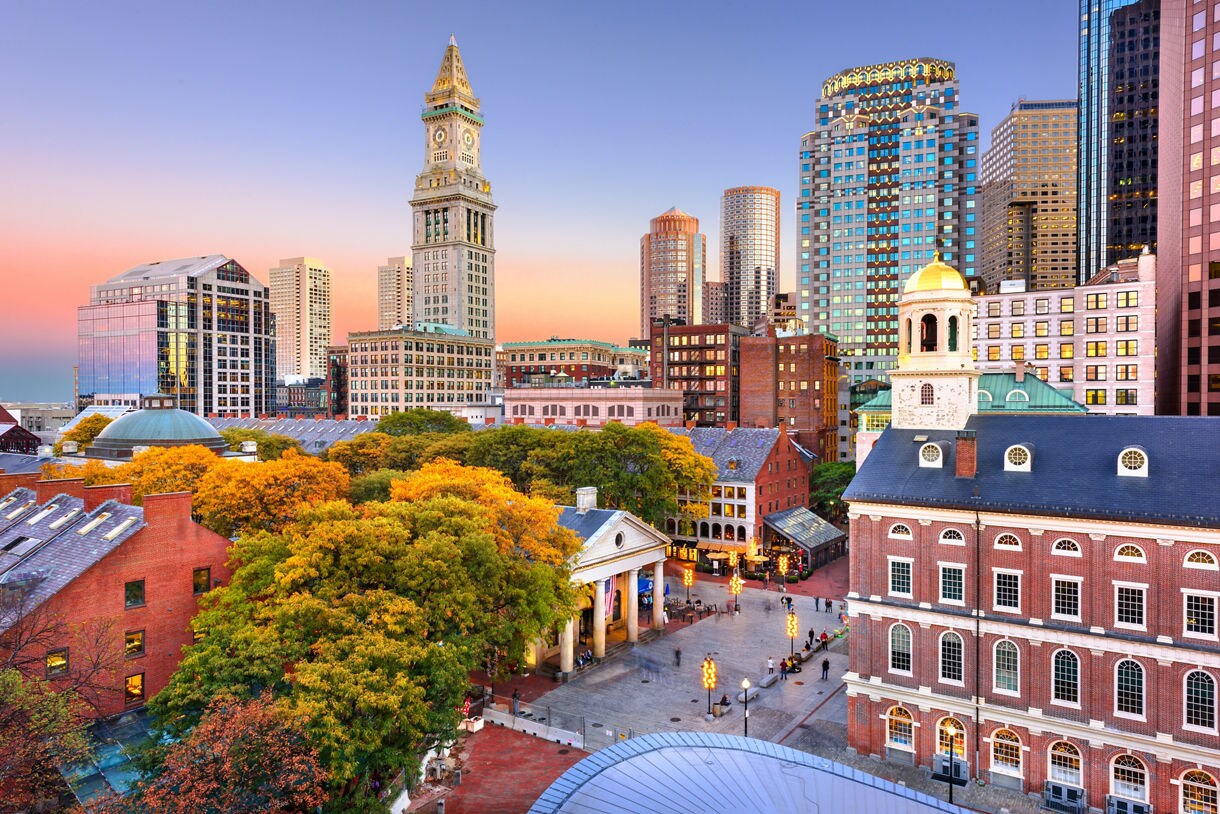 Aerial view of downtown Boston at sunset with colorful fall trees, historic brick buildings, Faneuil Hall and modern skyscrapers glowing in warm evening light.