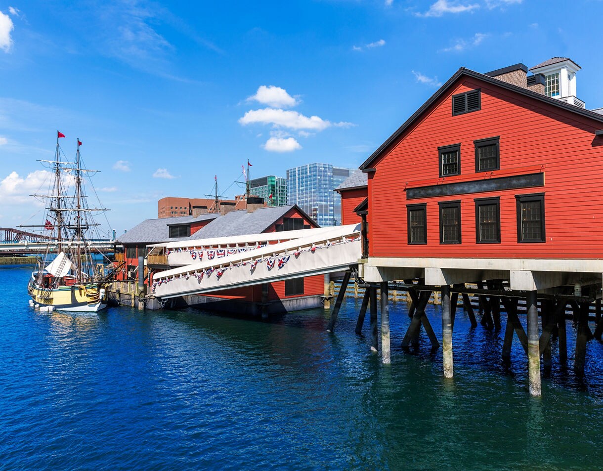 Red waterfront buildings on stilts and a docked historic tall ship on a sunny day at Boston Harbor, with modern city buildings rising in the background.