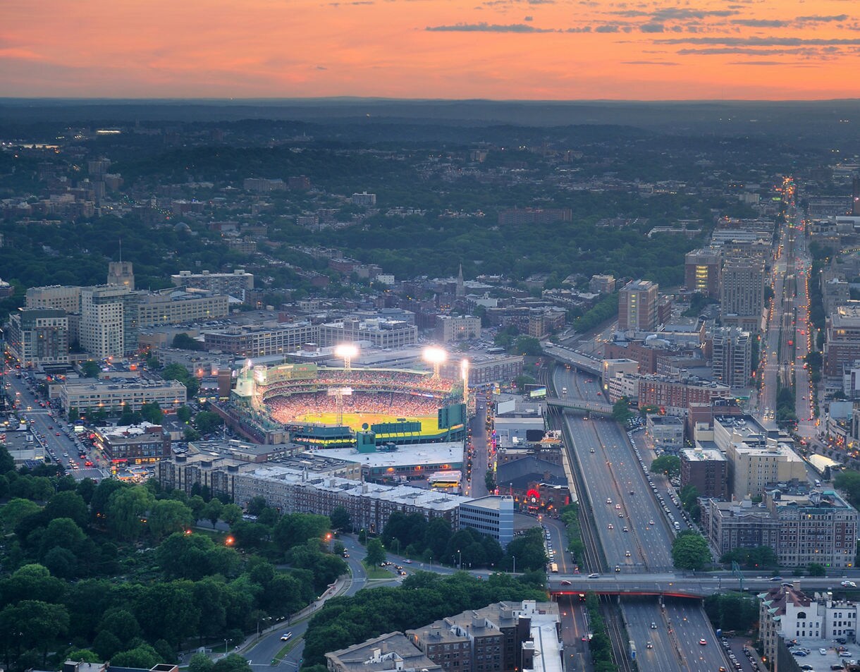 Aerial view of Fenway Park illuminated during an evening game, surrounded by city streets, traffic and dense urban neighborhoods at sunset.
