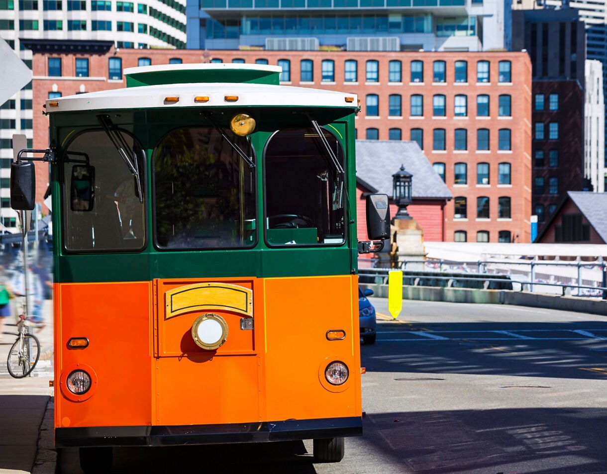 Bright orange and green Boston trolley parked along a street with brick buildings and modern high-rises in the background.