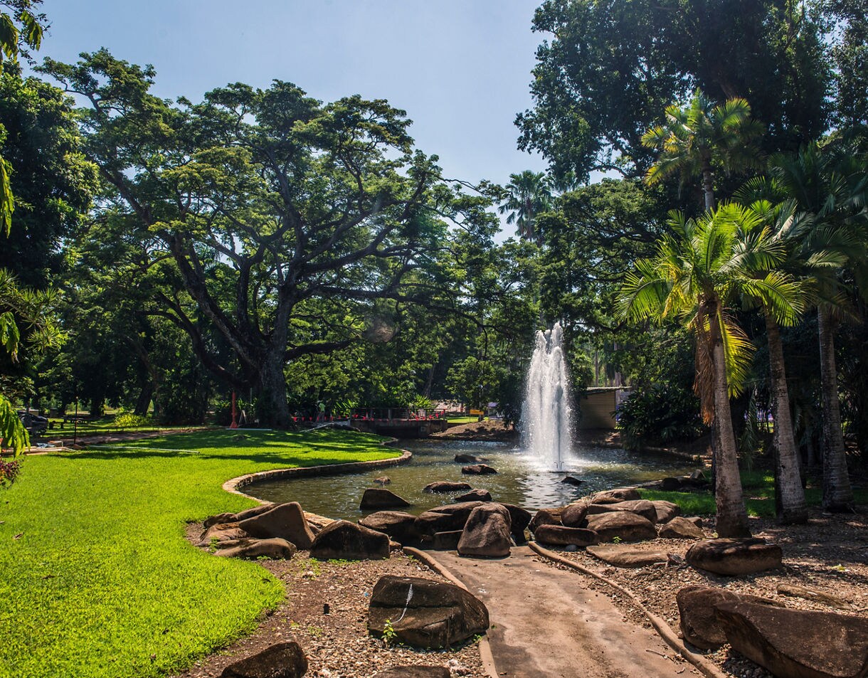 Fountain spraying water in a pond surrounded by palm trees, tropical plants and shaded walking paths in Darwin’s Botanic Gardens.