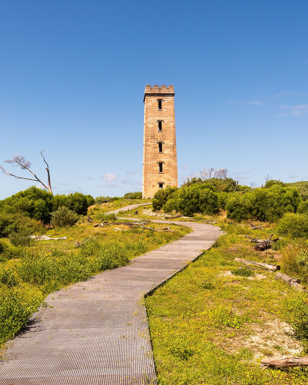 Tall sandstone tower rising above green coastal scrub, with a boardwalk path leading toward it under a bright blue sky.
