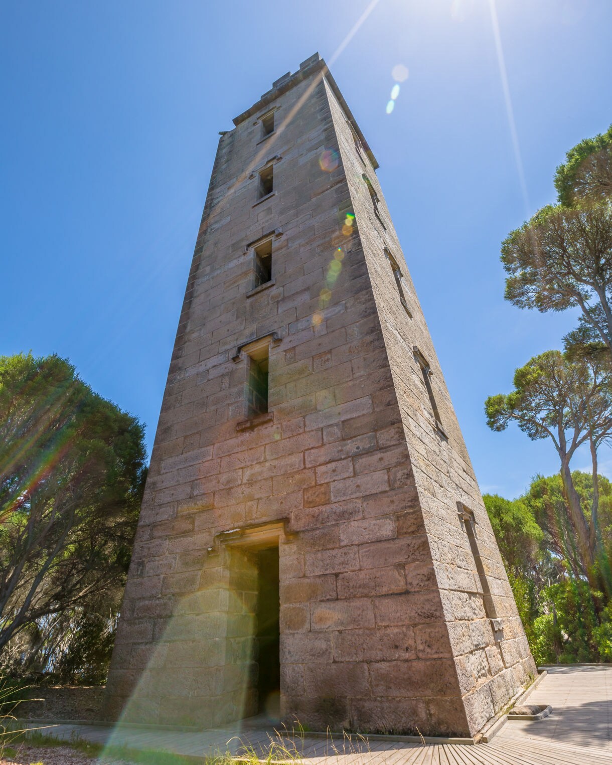 A tall, narrow stone tower photographed from a low angle, with bright sun flares overhead and surrounding trees framing the scene.