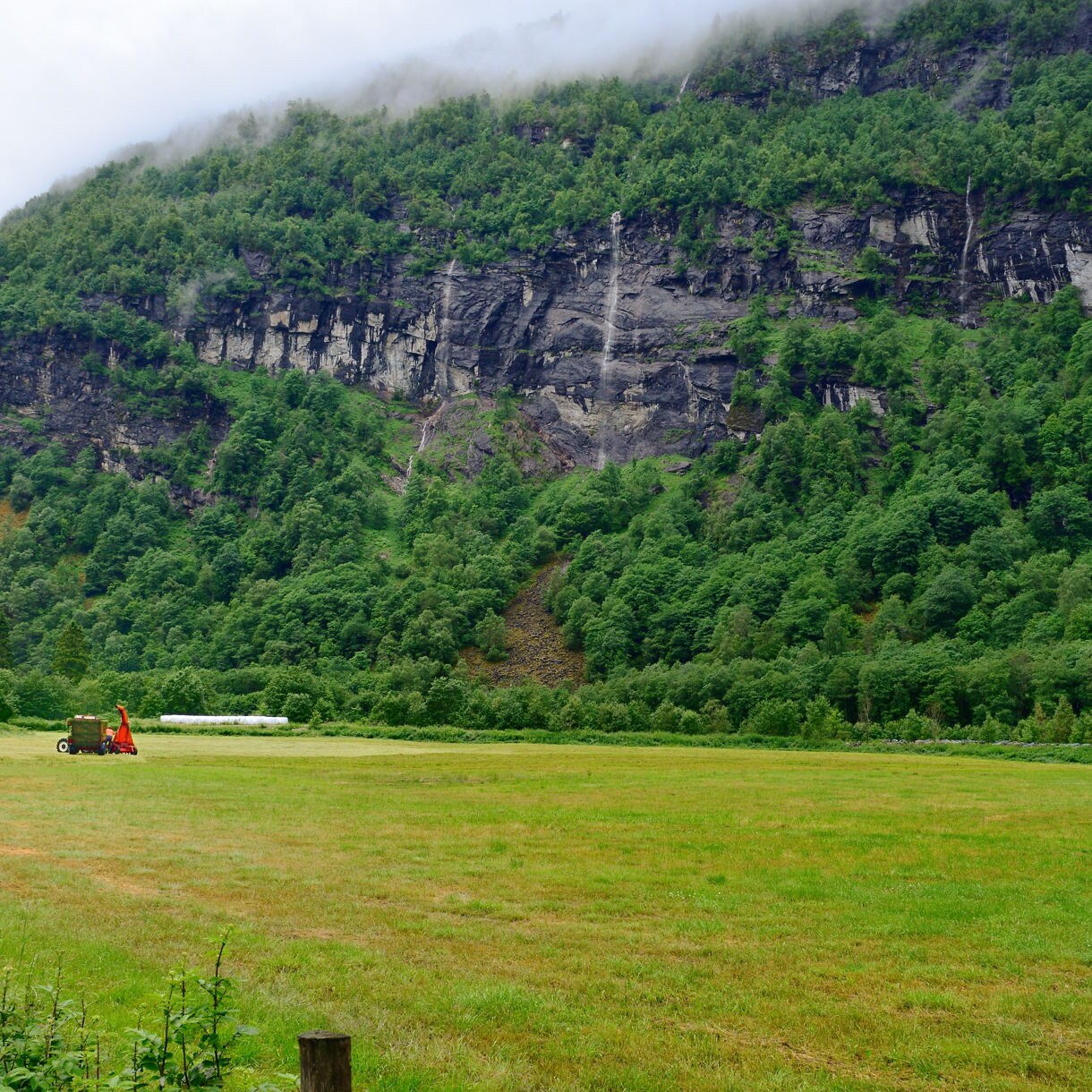 Farmland with a tractor and small shed in Breheimen National Park, Norway, set against steep forested cliffs with thin waterfalls cascading down.
