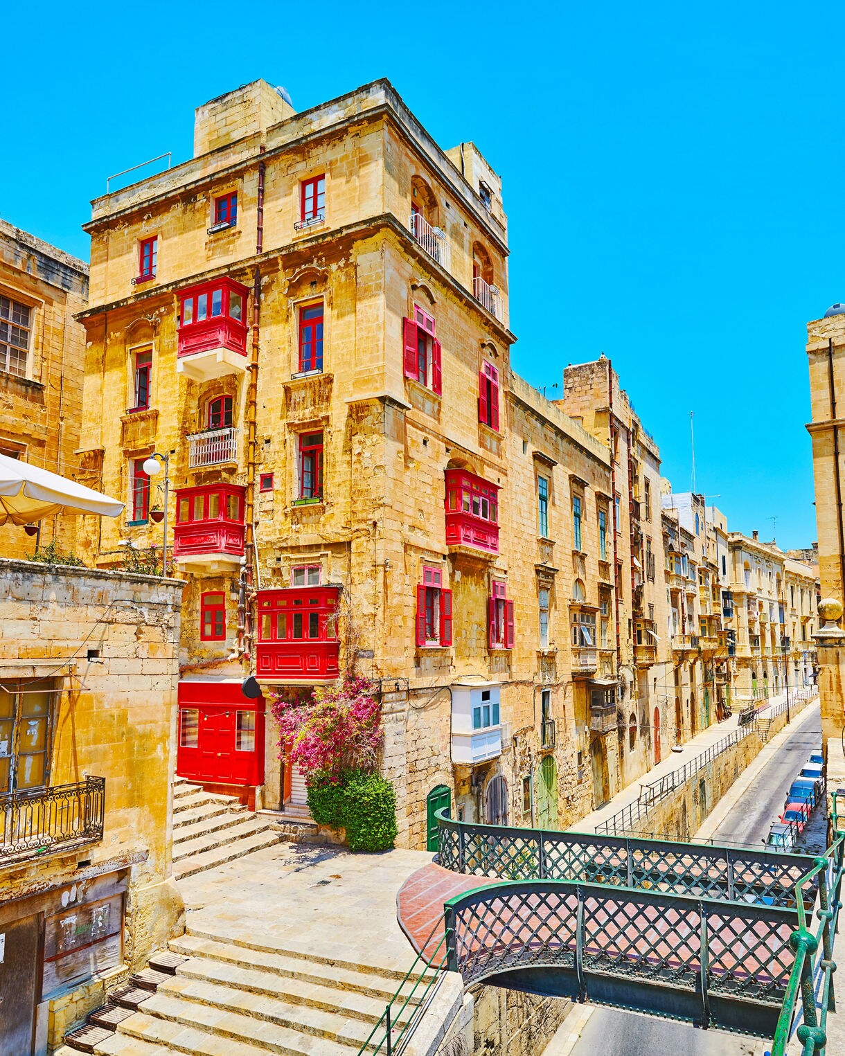 Historic limestone buildings in Valletta, Malta, featuring traditional red wooden balconies, arched doorways and a small bridge crossing a narrow street.