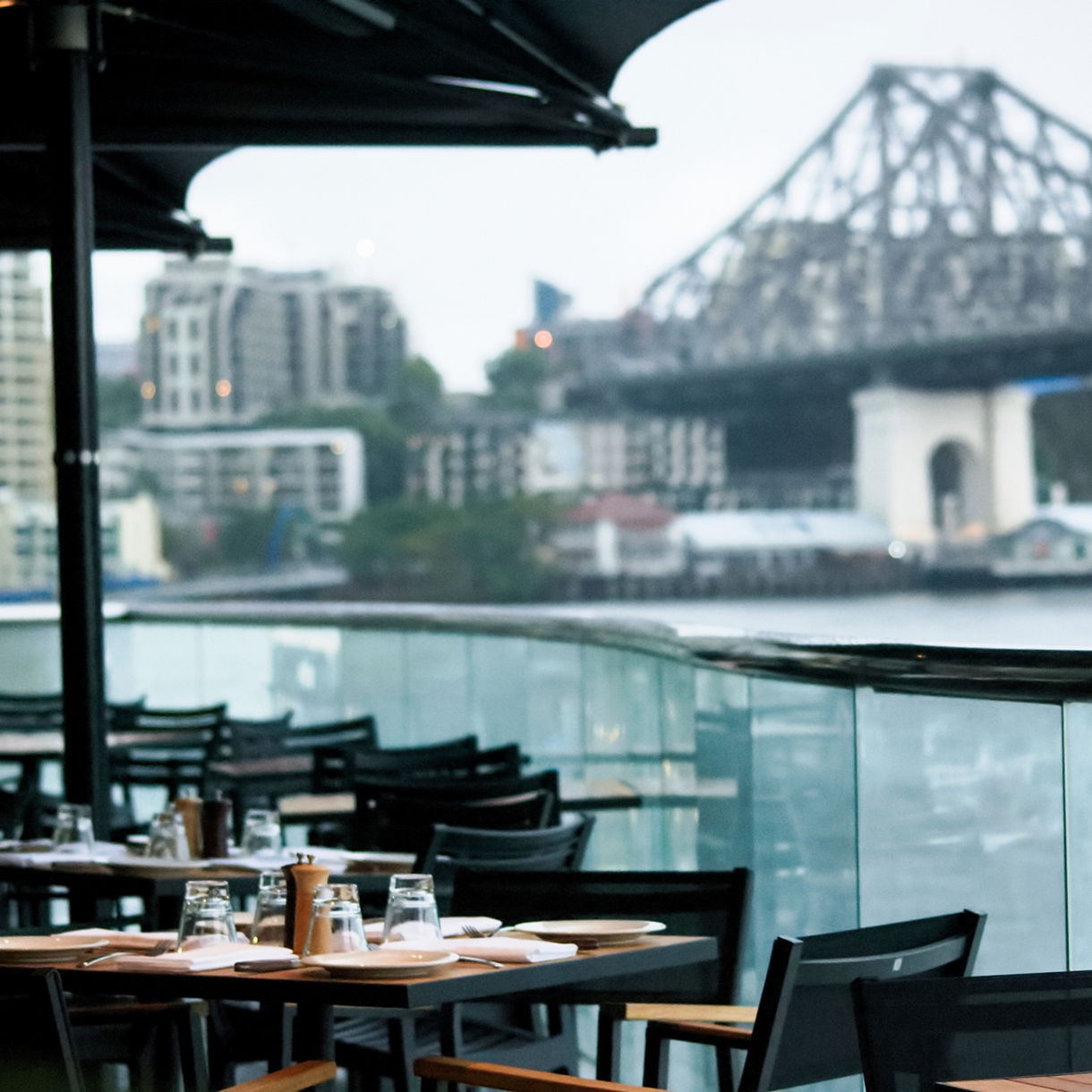 An outdoor cafe seating area with set tables and black chairs overlooking the Brisbane River, with the iconic steel structure of the Story Bridge visible in the blurred background.