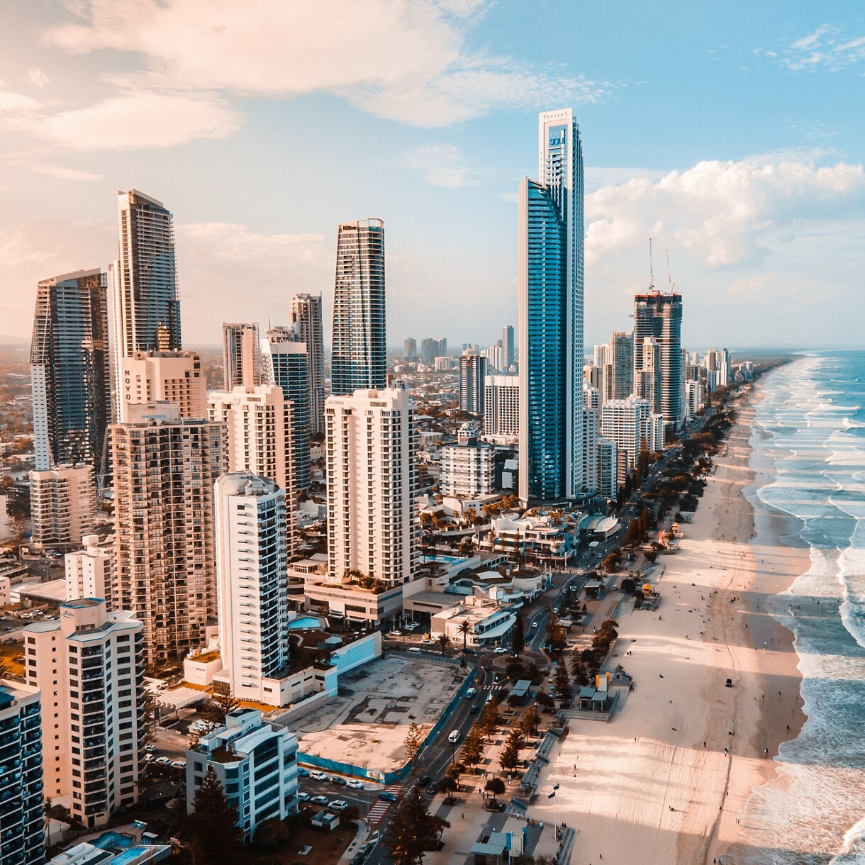 Aerial view of Australia's Gold Coast showing a densely packed strip of modern skyscrapers lining a broad sandy beach where white waves crash into the shore under a blue and pink-tinged sky.