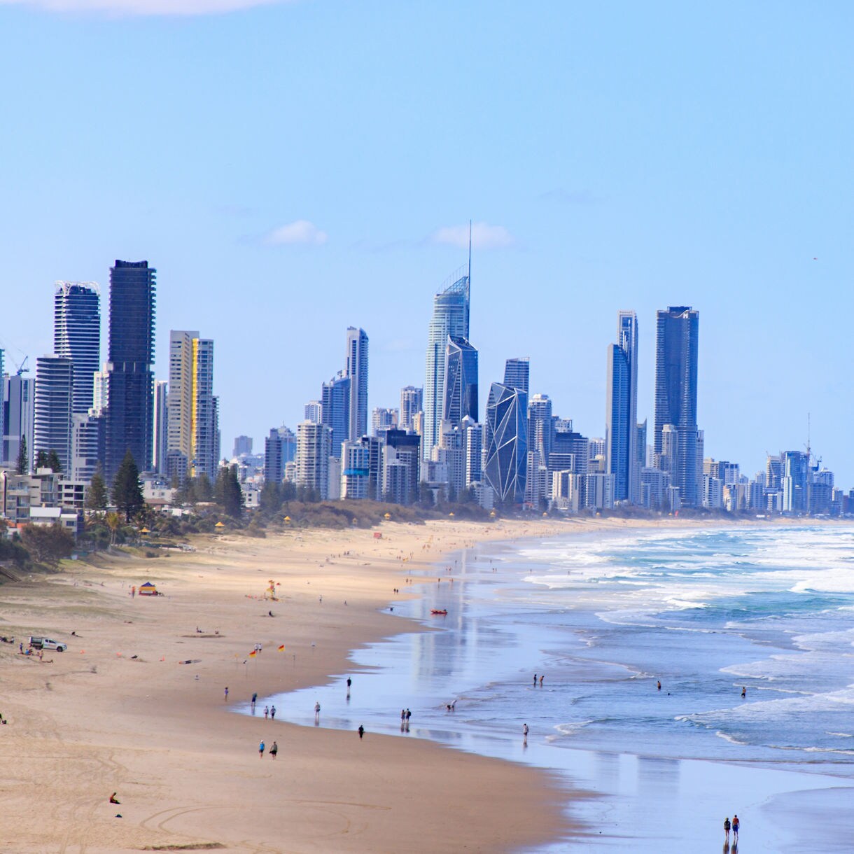 View of the Gold Coast Australia skyline showing numerous modern high-rise buildings towering above a wide reflective beach with ocean waves rolling in and scattered people walking along the wet sand.