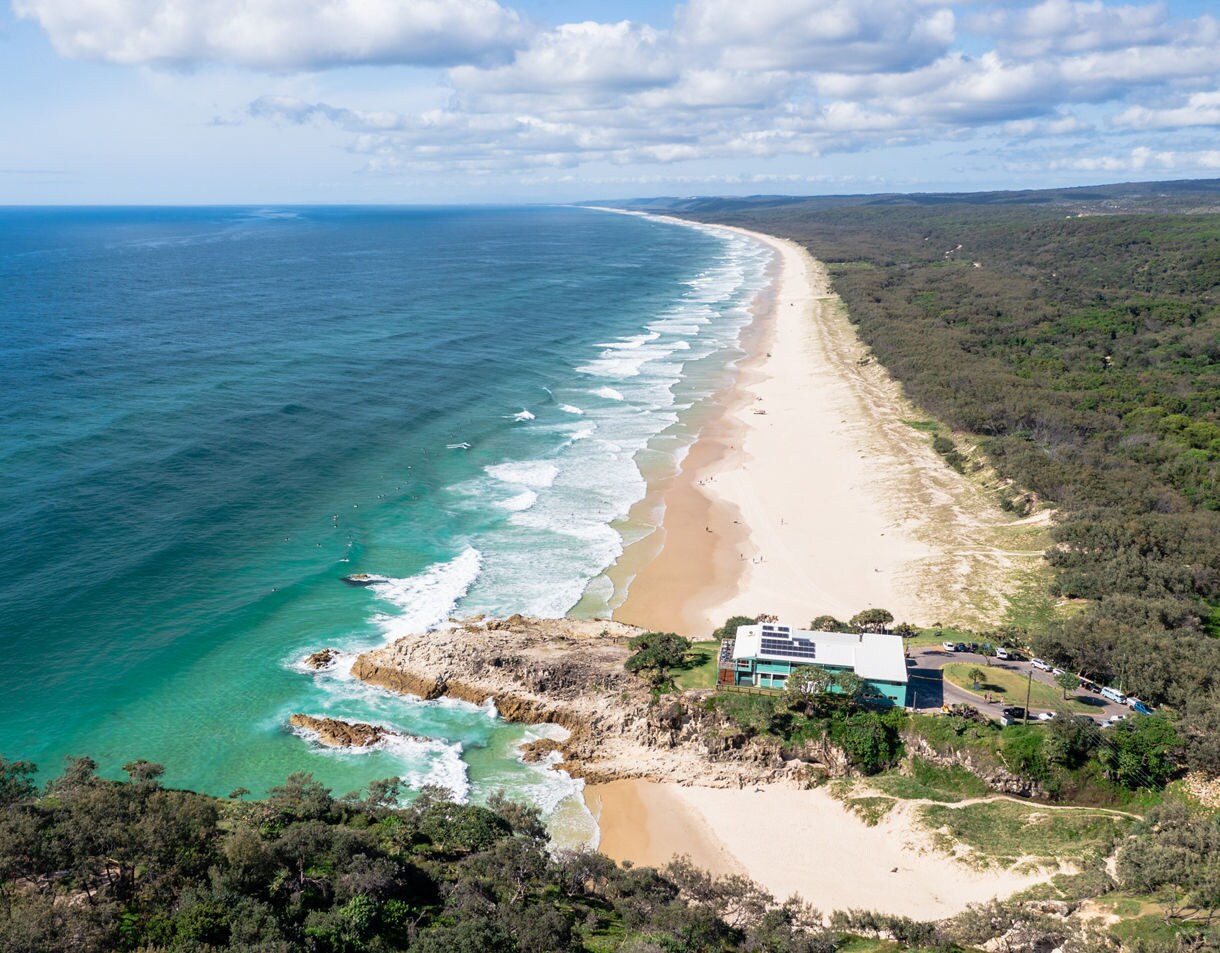Aerial view of North Stradbroke Island featuring a long expansive sandy beach bordered by dense green coastal vegetation, turquoise ocean water crashing onto the shore, and a small building near a rocky outcrop.