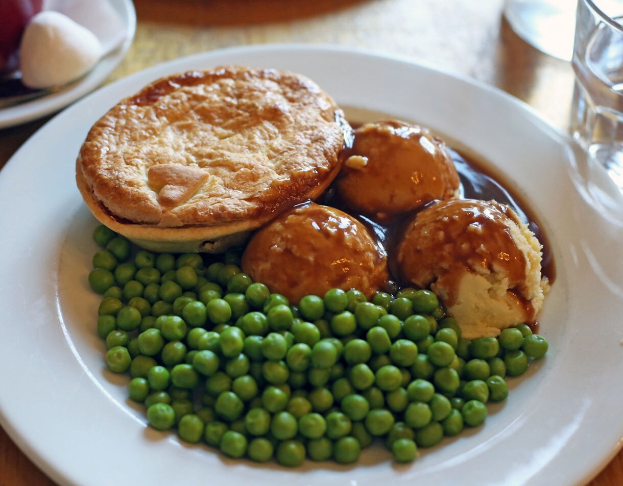 A classic Australian pub meal on a white plate featuring a golden-brown meat pie mashed potato balls covered in gravy and a generous serving of bright green peas.