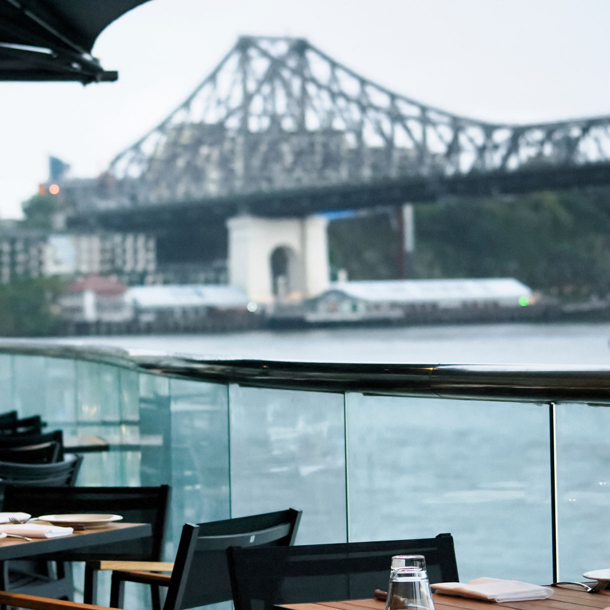 Set dining tables under black umbrellas on an outdoor riverfront patio, with the blurred steel frame of Brisbane’s Story Bridge spanning across the water in the background.