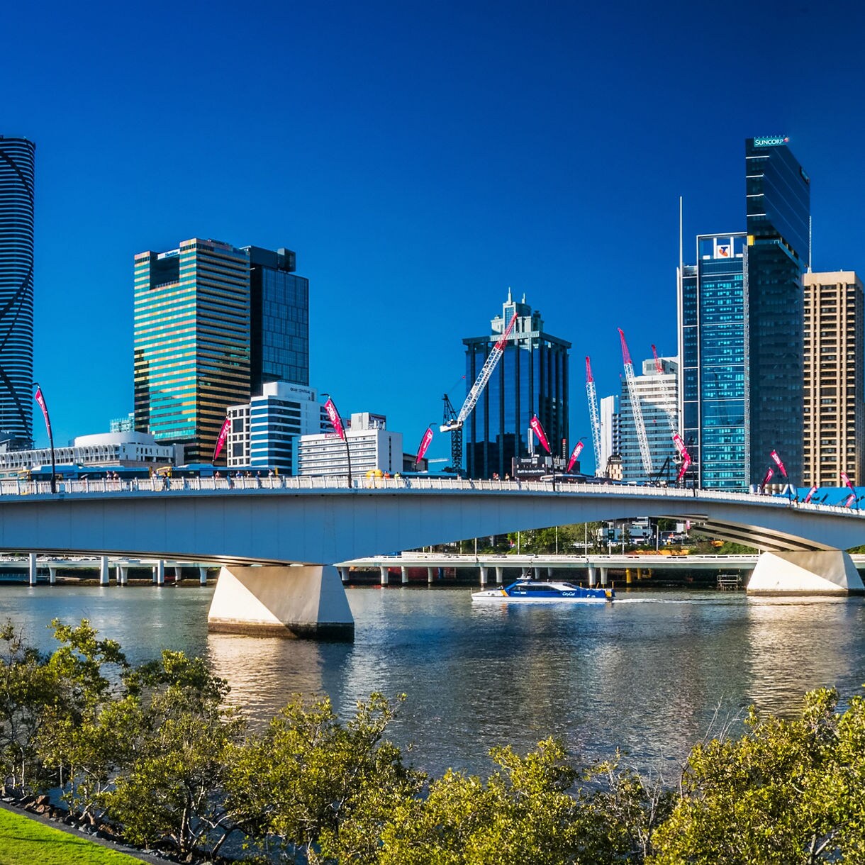 Panoramic view of the Brisbane Australia skyline featuring modern glass skyscrapers reflecting a clear blue sky, with a long white bridge spanning the Brisbane River and green trees in the foreground.