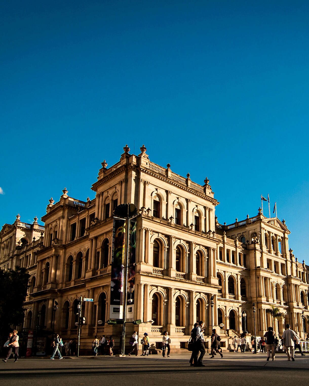The historic Treasury Building in Brisbane Australia a large sandstone structure with ornate architecture and arched windows viewed from the street level with pedestrians walking by under a brilliant blue sky.