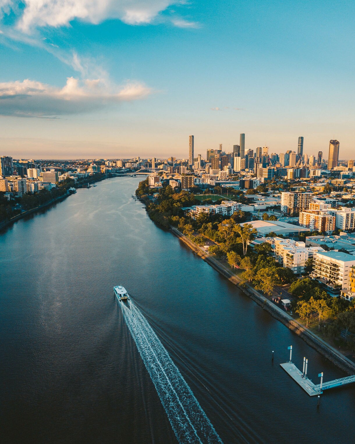 High-angle aerial view of the Brisbane River flowing toward the city skyline, with a wake visible behind a commuter ferry traveling on the dark blue water, all illuminated by warm late afternoon sun.