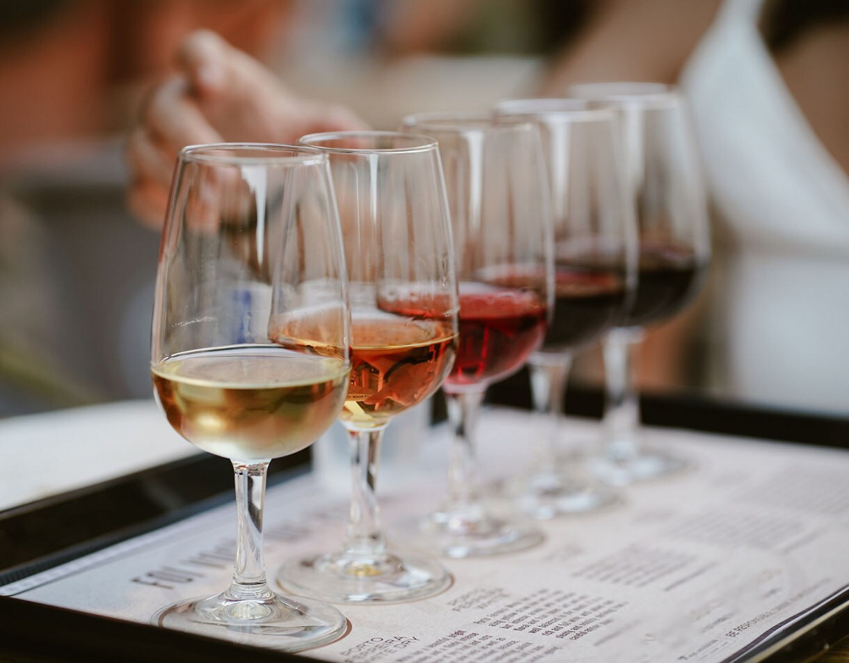 A close-up shot of a wine tasting flight featuring six glasses of wine ranging from white to amber to deep red, arranged in a line on a tasting menu placed on a black serving tray.