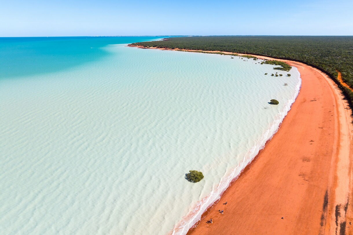 Aerial view of a long red-sand beach meeting pale turquoise water, with small mangroves scattered near the shore.