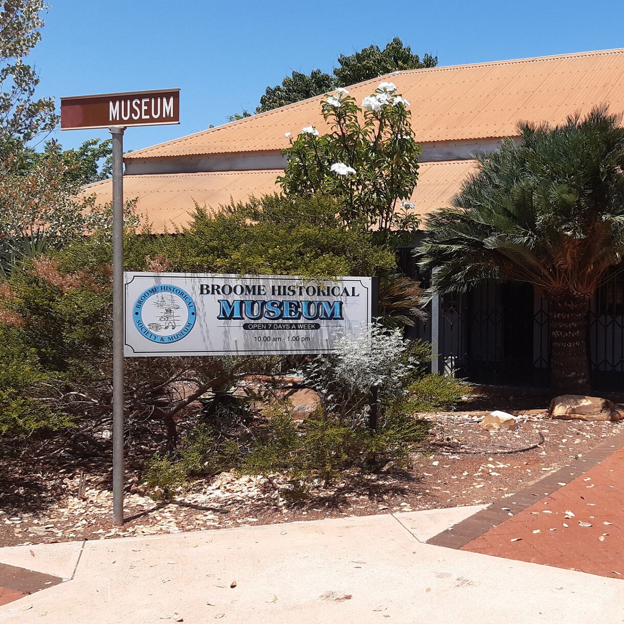 Entrance of the Broome Historical Museum with a sign, tropical plants and a tan roofed building on a sunny day.